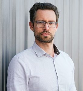 Nicolas Bengsch, Geschäftsführer der HNB Nordbleche GmbH, steht mit einem grauen Hemd und einer schwarzen Brille vor einem grauen Trapezblech Hintergrund
